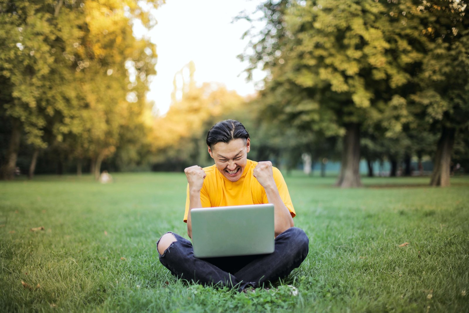 man sat down on grass with laptop with hands in a fist. He seems happy.